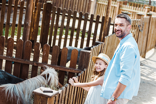 cheerful child and man smiling while standing near pony in zoo