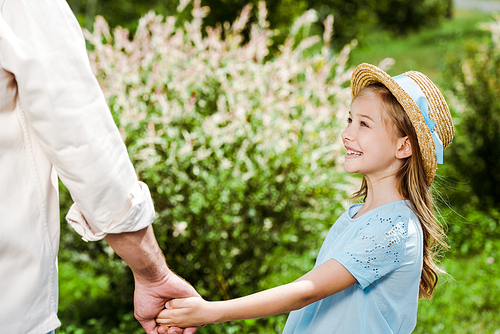 cropped view of father holding hands with cute and happy daughter in straw hat