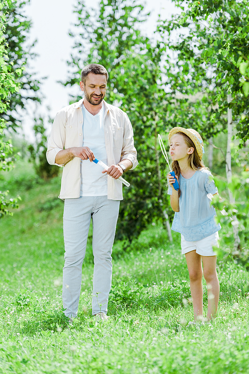 selective focus of cheerful father standing near daughter blowing soap bubbles near trees