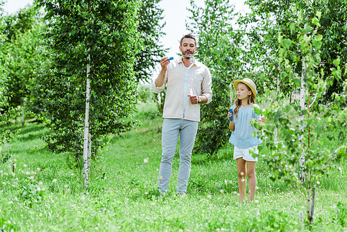 selective focus of father and daughter blowing soap bubbles near trees