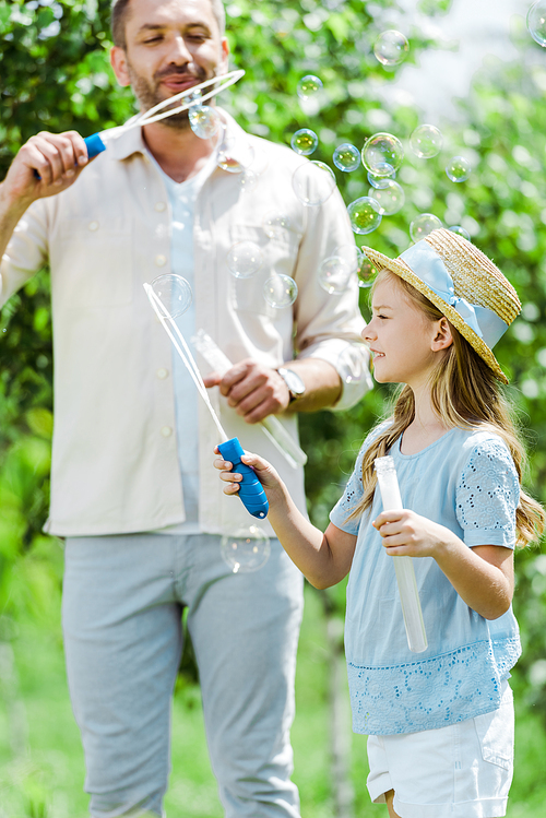 selective focus of cute daughter in straw hat holding bubble wand  near father blowing soap bubbles
