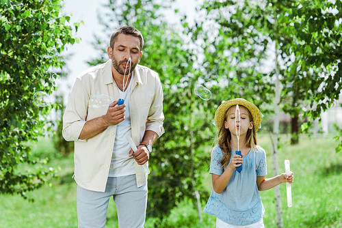 handsome man and daughter in straw hat blowing soap bubbles near trees