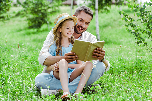 cheerful father and daughter sitting on grass and reading book
