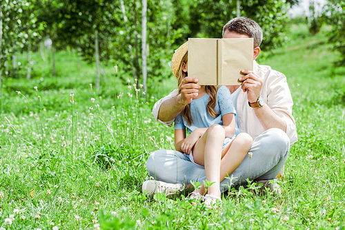 father and daughter covering faces while holding book and sitting on grass