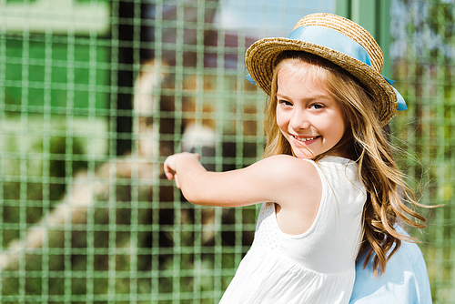 happy kid in straw hat  and gesturing near cage in zoo