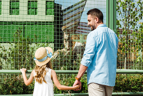 cheerful father looking at daughter while holding hands near cage in zoo