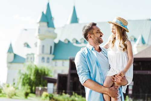 cheerful father holding in arms cute daughter in straw hat near castle