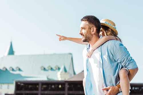 cheerful man piggybacking daughter pointing with finger near building