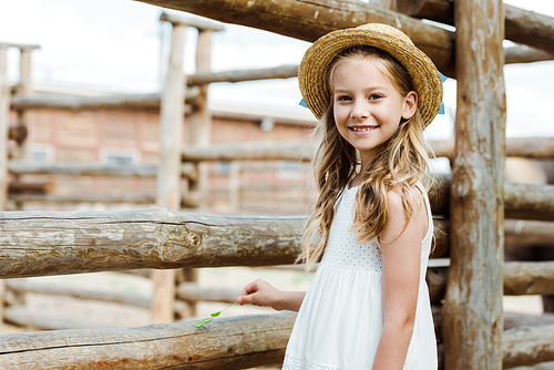 happy kid in straw hat standing in white dress near fence in zoo