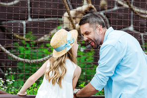 kid in straw hat looking at father showing tongue near cage with wild animal in zoo