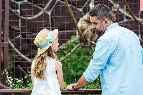kid in straw hat looking at upset father near cage with wild animal in zoo