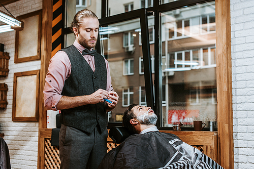 handsome barber holding razor near bearded man with shaving cream on face
