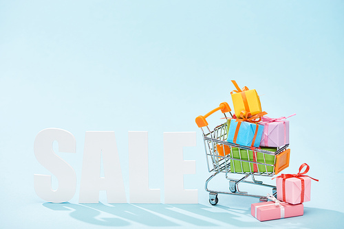 white sale lettering and festive gift boxes with bows in shopping cart on blue background