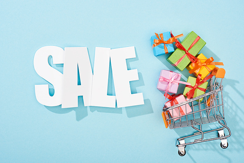 top view of white sale lettering and colorful gift boxes in shopping cart on blue background