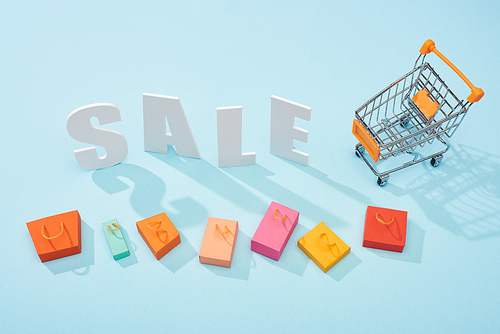 top view of white sale lettering near shopping trolley and colorful shopping bags on blue background
