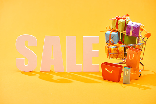 white sale lettering with shopping bags and festive gifts in shopping cart on bright orange background
