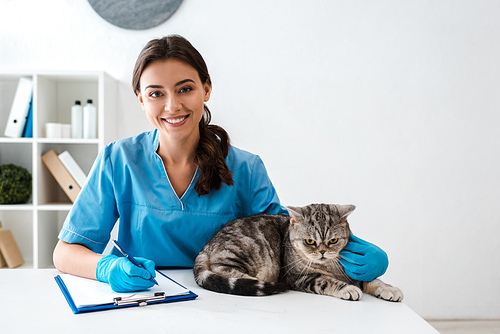 young veterinarian smiling at camera while writing on clipboard near tabby scottish straight cat