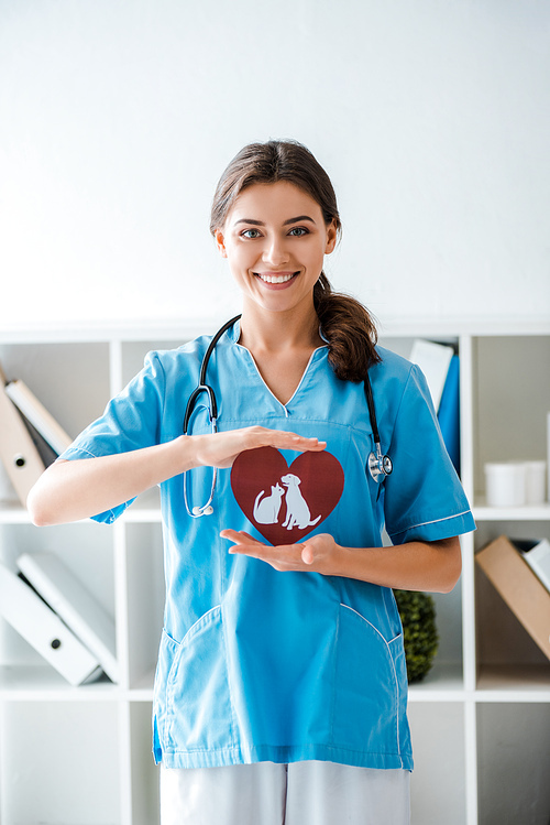 attractive, positive veterinarian smiling at camera while presenting paper cut heart with dog and cat symbols