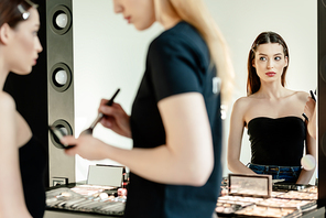 selective focus of beautiful model looking at mirror near makeup artist