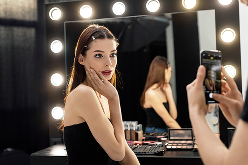 selective focus of makeup artist taking photo of beautiful model while using smartphone