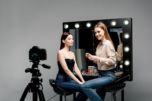 selective focus of happy makeup artist holding cosmetic brush and lip gloss near model and digital camera isolated on grey