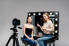 selective focus of happy makeup artist holding lip gloss near model and digital camera isolated on grey