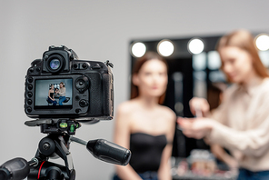 selective focus of digital camera with makeup artist holding lip gloss near model on screen isolated on grey