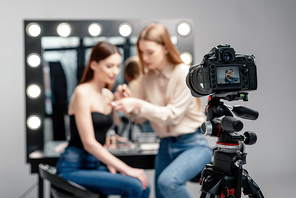 selective focus of digital camera near makeup artist holding lip gloss near model isolated on grey