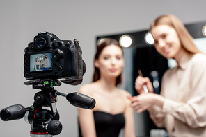 selective focus of digital camera with cheerful makeup artist holding lip gloss near model on screen isolated on grey