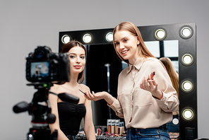 selective focus of smiling makeup artist holding lip gloss near model and looking at digital camera isolated on grey