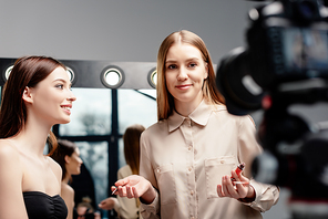selective focus of makeup artist holding lip gloss near happy model and looking at digital camera isolated on grey