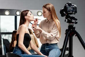 selective focus of makeup artist applying lip gloss on lips of beautiful model near digital camera isolated on grey