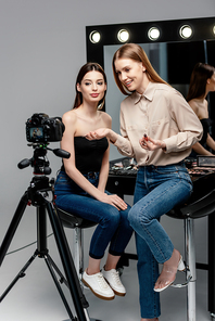 selective focus of happy makeup artist holding lip gloss near beautiful model and digital camera on grey