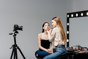 cheerful makeup artist applying lip gloss on lips of attractive model and looking at digital camera on grey