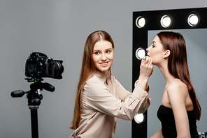 selective focus of happy makeup artist applying lip gloss on lips of attractive model and looking at digital camera isolated on grey