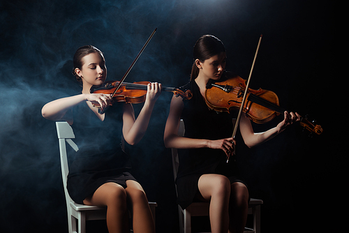 attractive musicians playing on violins on dark stage with smoke