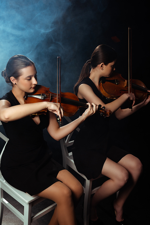 attractive professional musicians playing on violins on dark stage with smoke