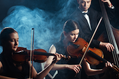 trio of musicians playing on violins and contrabass on dark stage with smoke