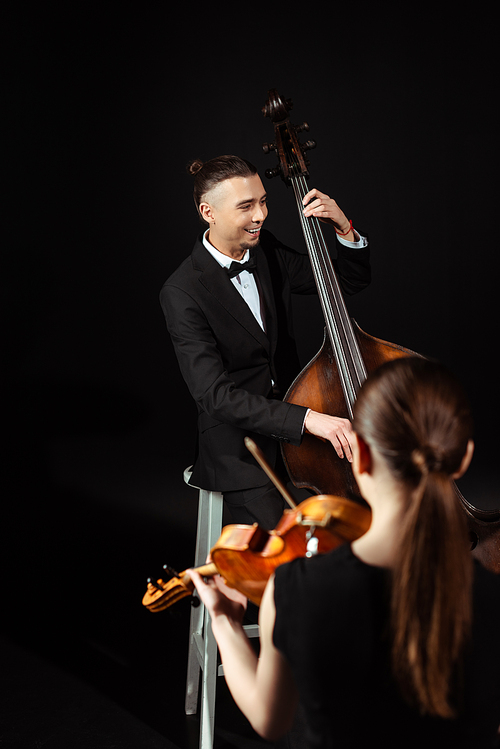 cheerful professional musicians playing on violin and contrabass on dark stage