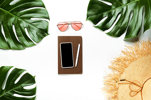 top view of green palm leaves, straw hat, sunglasses and notepad with smartphone on white background