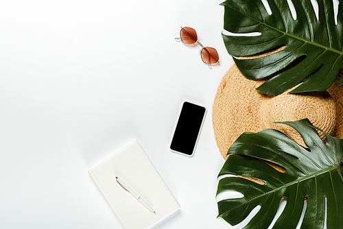 top view of green palm leaves, straw hat, sunglasses and notepad with smartphone on white background