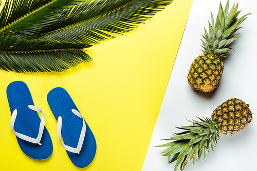 top view of green palm leaves, pineapples and blue flip flops on white and yellow background