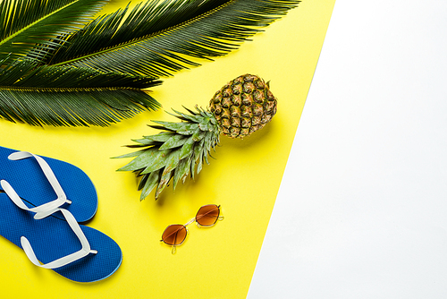 top view of green palm leaves, pineapple, sunglasses and blue flip flops on white and yellow background