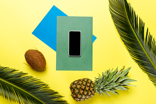 top view of green palm leaves, smartphone, coconut, pineapple on colorful background