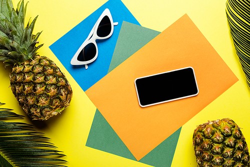 top view of green palm leaves, sunglasses, smartphone and ripe pineapples on colorful background