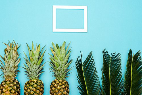 top view of green palm leaves and ripe pineapples near square empty frame on blue background