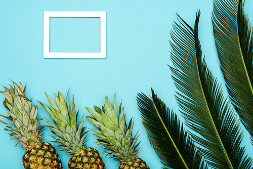 top view of green palm leaves and ripe pineapples near square empty frame on blue background