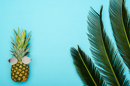 top view of green palm leaves and ripe pineapple with sunglasses on blue background