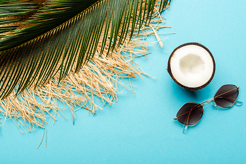 top view of green palm leaf, coconut half, sunglasses and straw hat on blue background