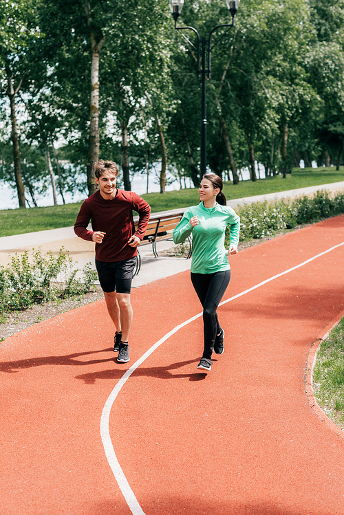 Beautiful woman smiling while jogging near boyfriend on running track in park